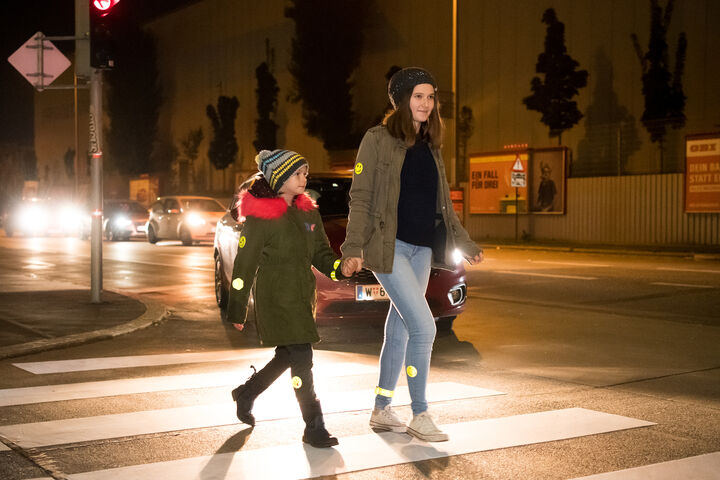 Ein Kind uns eine erwachsene Person überqueren in der Dunkelheit einen Zebrastreifen, im Hintergrund sieht man ein Auto.