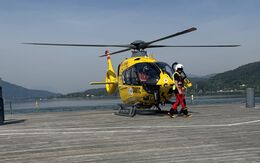 Christophorus-Hubschrauber steht auf einem Steg am See. Vor dem Hubschrauber geht ein Crew-Mitglied.