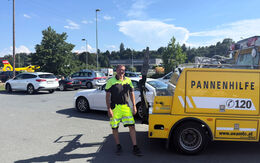 ÖAMTC Pannenhelfer steht auf Parkplatz neben gelben Pannenhilfeauto. Im Hintergrund ist ein Christophorus-Hubschrauber zu sehen.