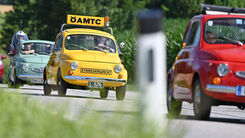 Kleine bunte Autos fahren auf einer Landstraße. Eines der Autos ist gelb und hat ein ÖAMTC-Schild auf dem Dach.