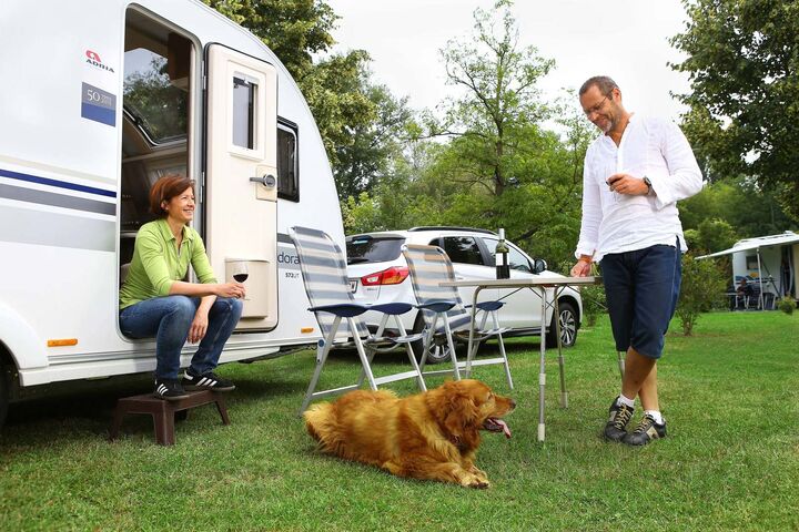 Auf einem Campingplatz sitzt eine Frau vor einem Wohnwagen, während ein Mann mit einem Hund auf der Wiese spielt.