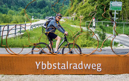 Das Bild zeigt den auto touring-Redakteur, der auf dem Ybbstal-Fahrradweg unterwegs ist und vor einem großen orangefarbenen Sockel posiert, auf dem "Ybbstalradweg" steht. Auf dem Sockel befindet sich auch eine Metallskulptur von zwei Radfahrern. Im Hintergrund sieht man eine Straße in grüner Landschaft mit Bäumen und Hügeln.