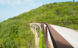 Ein Radfahrer überquert ein großes Viadukt in einer mit Bäumen bedeckten hügeligen Landschaft. Der Radweg ist Teil einer stillgelegten Bahnstrecke von Triest hinauf in den Karst.