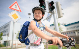 Ein Junge fährt auf einem Fahrrad. Er trägt einen Helm und eine blaue Schultasche am Rücken.