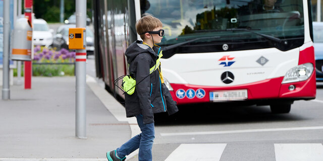 Ein Kind mit einer Blickbewegungsbrille überquert die Straße. Im Hintergrund sieht man einen Autobus.