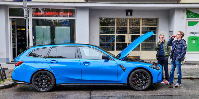 Ein blauer BMW M3 steht mit geöffneter Motorhaube auf einem Parkplatz, zwei Personen stehen neben dem Auto.