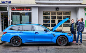 Ein blauer BMW M3 steht mit geöffneter Motorhaube auf einem Parkplatz, zwei Personen stehen neben dem Auto.