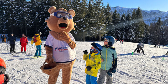 Maskottchen Kasbär Bruno im Bärenkostüm auf der Skipiste am Kasberg. Zwei Kinder stehen gebannt vor ihm. 