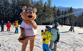 Maskottchen Kasbär Bruno im Bärenkostüm auf der Skipiste am Kasberg. Zwei Kinder stehen gebannt vor ihm. 