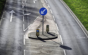 Eine Straße an der Linksverkehr herrscht. Eine Straßenschild auf dem ein Pfeil abgebildet ist, der nach links unten zeigt.