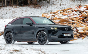 Ein schwarz-roter Mazda MX-30 steht in einer verschneiten Winterlandschaft vor einem Haufen Holz.