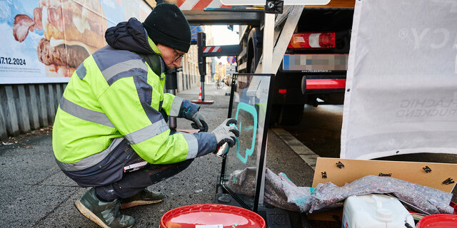 Graffiti-Künster sizetwo sprüht auf eine Glasplatte etwas auf. Er hockerlt dabei neben einem LKW am Gehsteig.