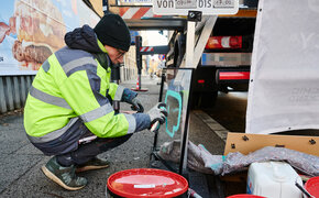 Graffiti-Künster sizetwo sprüht auf eine Glasplatte etwas auf. Er hockerlt dabei neben einem LKW am Gehsteig.