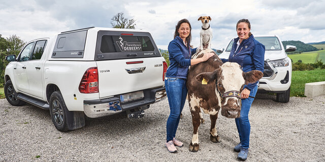 Zwei Frauen stehen mit einer Kuh, auf der ein Hund sitzt vor zwei Autos. Sie sind Tierärztinnen.