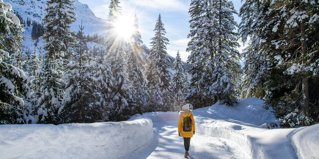 Eine Frau in einem gelben Anorak spaziert durch einen verschneiten Wald. 