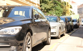 Parkende Autos stehen am Straßenrand einer Kurzparkzone im Sonnenschein. Im Hintergrund sind Mietshäuser, ein großer Baum und ein Kran sichtbar.