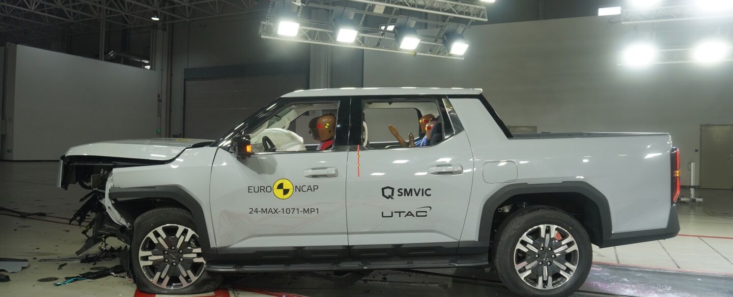 Crashtest von einem weißen großen Pickup. Die Motorhaube ist stark eingedrückt und kaputt. Das Fahrzeug steht in einer Testhalle.