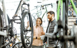 Ein junger Mann und eine junge Frau sehen sich in einem Fahrradgeschäft ein Fahrrad an. Beide lachen und der Mann gibt das Daumen-hoch-Zeichen.